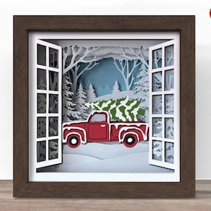 May include: A framed paper art piece depicting a winter scene. A red pickup truck carries a Christmas tree through a snowy landscape, viewed through a window. The frame is brown, and the window is white.