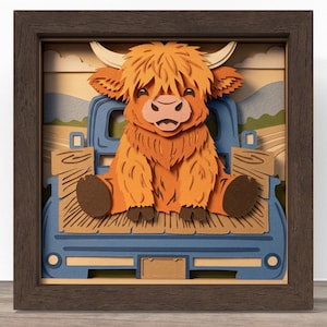 Könnte beinhalten: Gerahmtes 3D-Papierkunstwerk mit einem Highland-Rind, das in einem blauen Pickup-Truck sitzt. Das Rind ist orange und braun, mit Hörnern und einem flauschigen Fell. Der Hintergrund zeigt eine ländliche Landschaft mit Feldern und Bergen.