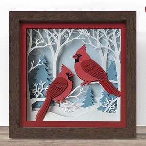 May include: A square shadow box frame with a dark wood finish containing a layered paper art scene. The artwork features two red cardinal birds perched on white branches against a backdrop of blue pine trees, white trees, and snowflakes.