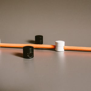 Könnte beinhalten: Vier Kabelclips, zwei schwarze und zwei weiße, sind an einem orangefarbenen Kabel befestigt. Die Kabelclips sind so konzipiert, dass sie Kabel an Ort und Stelle halten.