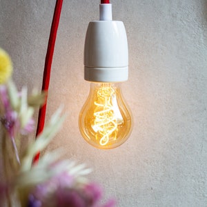Könnte beinhalten: Eine weiße Keramik-Lampenfassung mit einem roten Kabel, das von der Decke hängt. Eine Glühbirne mit Spiralfilament ist in die Fassung geschraubt und leuchtet.