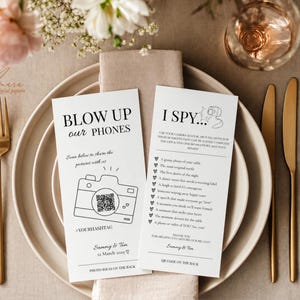 May include: Two white rectangular wedding game cards with black text and line art. One card features a camera illustration with a QR code and the text BLOW UP our PHONES. The other card is titled I SPY and lists photo prompts for guests.