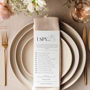 May include: A white rectangular I Spy wedding game card with black text and a camera icon, placed on a beige linen napkin atop stacked ceramic plates. The card lists photo prompts and includes the names Sammy & Tim at the bottom.