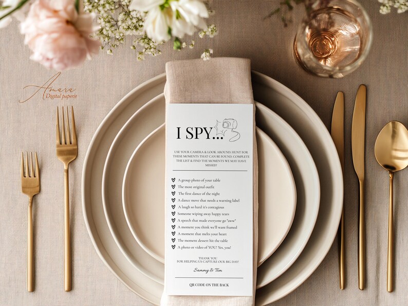 May include: A white rectangular I Spy wedding game card with black text and a camera icon, placed on a beige linen napkin atop stacked ceramic plates. The card lists photo prompts and includes the names Sammy & Tim at the bottom.