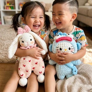 May include: Two children smiling and holding handmade crochet dolls. One doll is a white bunny with a pink bow and pink accents. The other is a blue doll with a red bow and a cat face.