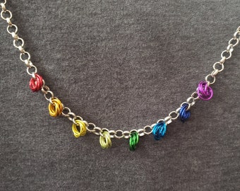 Puede incluir: Un collar de cadena plateada con anillos de alambre de colores arcoíris. Los anillos están en el orden del arcoíris, rojo, naranja, amarillo, verde, azul, índigo y violeta.