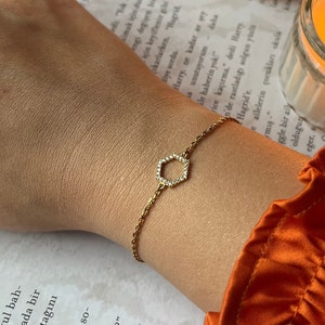 May include: A gold chain bracelet with a small, geometric, diamond-shaped pendant. The bracelet is on a person's wrist, and the background is a book and a lit candle.