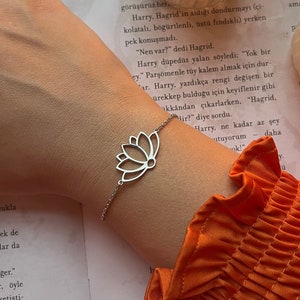May include: A silver bracelet with a delicate lotus flower charm. The bracelet is on a person's wrist, and the background is a book with orange text.