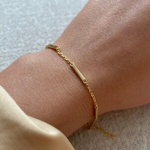 May include: A gold chain bracelet with three small, rectangular gold bars attached to the chain.