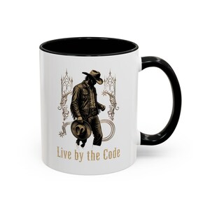Peut inclure: Mug en céramique blanche avec intérieur et anse noirs. L'illustration représente un cowboy avec le texte "Live by the Code" en lettres dorées. Le cowboy porte un chapeau et une veste.