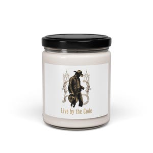 Peut inclure: Un pot de bougie blanc avec un couvercle noir. L'étiquette présente une illustration sépia d'un cow-boy tenant un chapeau, avec le texte "Live by the Code" en dessous. La bougie est probablement parfumée.