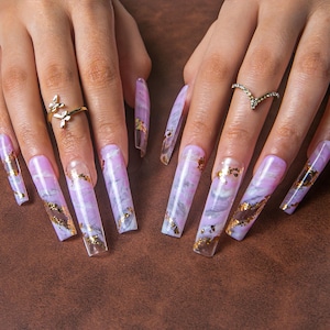 May include: Long, square-shaped artificial nails with a marble design in shades of lavender and gray, accented with gold foil. The nails are adorned with gold rings, including a butterfly and a chevron design.