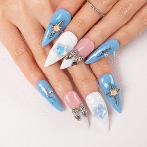 May include: A hand displaying long, pointed fingernails with a variety of nail art. The nails are painted in light blue, white, and pink. Some nails feature silver embellishments like bows, stars, and cross-like designs.