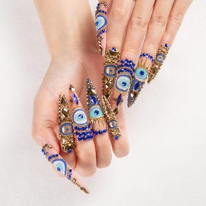 May include: A hand displaying long, pointed fingernails adorned with intricate nail art. The nails feature a combination of gold, blue, and turquoise embellishments, including eye-shaped designs and sparkling gems. The overall effect is glamorous and eye-catching.