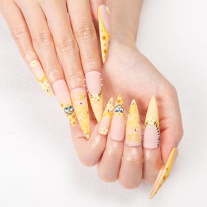 May include: A set of long, pointed artificial nails. The nails feature a light yellow and nude color scheme with cartoon character designs, floral accents, and rhinestones. The design includes polka dots and swirls.
