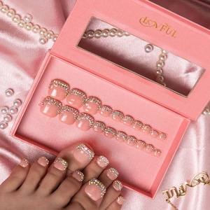 May include: A set of press-on toenails in a pink box with the word "LOVFUL" in gold lettering. The nails are a light pink with a sparkling rhinestone border. A foot with similar nails is also visible.