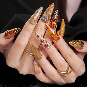 May include: A close-up of a hand displaying a set of long, ornate, and bejeweled artificial nails. The nails feature various designs, including gold, brown, orange, and white, with embellishments like gems and textured patterns. Gold rings adorn the fingers.