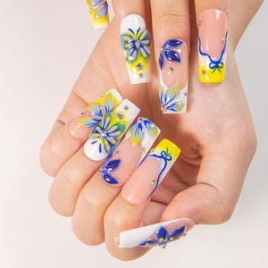 May include: A hand displaying long, artificial nails with floral designs. The nails feature a variety of colors, including white, yellow, blue, and pink, with intricate flower and leaf patterns. Some nails have rhinestones for added detail.