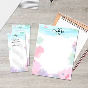 Op de afbeelding: Een set van twee notitieboekjes in aquarelstijl met een blauw en roze ontwerp. Het grotere notitieboekje heeft het woord "Notes" in cursief op de voorkant. Het kleinere notitieboekje heeft een lijst met items om te kopen, waaronder "drycleaner", "grocery store" en "pharmacy".