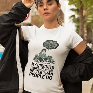 May include: White t-shirt with the slogan "MY CIRCUITS UNDERSTAND ME BETTER THAN PEOPLE DO." The graphic shows a person with headphones, circuit boards, and a thought bubble filled with circuit diagrams.