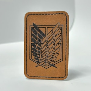 Könnte beinhalten: Ein rechteckiger, hellbrauner Lederaufnäher mit einem schwarzen Emblem, das stilisierte Flügel in einer Schildform zeigt. Der Aufnäher hat einen genähten Rand und ist wahrscheinlich ein dekorativer Artikel oder ein Accessoire.