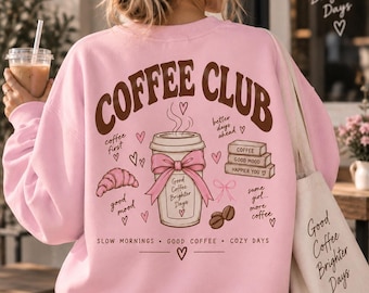 May include: A light pink crewneck sweatshirt featuring a brown graphic on the back that reads COFFEE CLUB. The design includes a coffee cup with a pink bow, a croissant, coffee beans, and stacked books with the text Good Coffee Brighter Days.