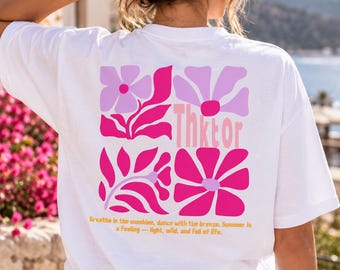 May include: A white t-shirt featuring a back graphic of four squares containing stylized pink and lavender flowers. The text "Thktor" is printed vertically in the center, and the phrase "Breathe in the sunshine, dance with the breeze. Summer is a feeling — light, wild, and full of life." is printed in yellow below the graphic.