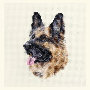 May include: A cross-stitch depiction of a German Shepherd dog's head in profile. The dog has a black nose, brown and black fur, and a pink tongue. The background is a light beige fabric, with the cross-stitch pattern clearly visible.
