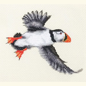 May include: Cross-stitch design of a puffin in flight. The bird has a white body, black wings and back, and a distinctive orange and yellow beak. Its feet are bright orange. The background is a light beige fabric.