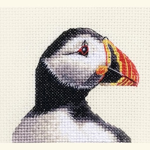 May include: Cross-stitch depiction of a puffin's head. The bird features a black and white body, a red eye, and a vibrant beak with red, yellow, and orange stripes. The background is a light beige fabric.