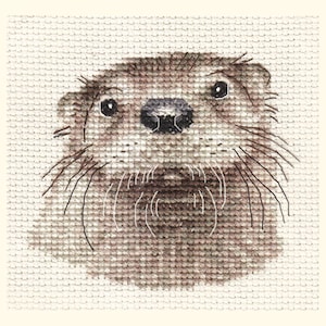 May include: Cross-stitch image of an otter's head. The otter is rendered in shades of brown and tan, with black eyes and long white whiskers. The background is a light beige. The cross-stitch technique gives the image a textured appearance.
