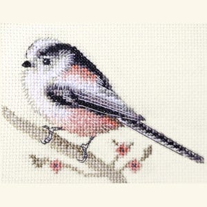May include: Cross-stitch embroidery of a long-tailed tit bird on a branch with pink flowers. The bird has a black and white head, grey wings, and a pinkish-brown body. The background is a light beige fabric.