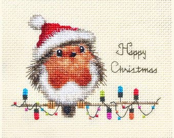 May include: Cross-stitch design of a bird wearing a Santa hat, perched on a string of Christmas lights. The bird has a red breast and is surrounded by a white border. The words "Happy Christmas" are stitched to the right.