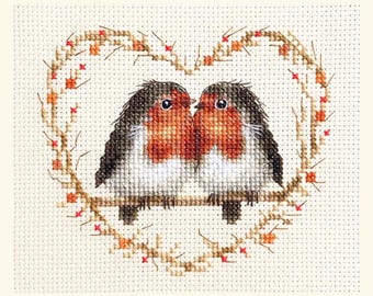 May include: Cross-stitch of two robins perched on a branch within a heart-shaped wreath. The birds have red breasts, white bellies, and gray and black wings. The wreath is made of brown twigs with small red accents, all on a cream-colored background.