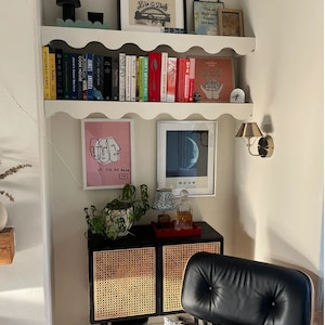 May include: A white shelf with scalloped edges, holding books, artwork, and a small clock. The shelf is mounted on a white wall. Below the shelf is a black cabinet with two doors, each with a woven rattan panel. A black leather chair is in the foreground.