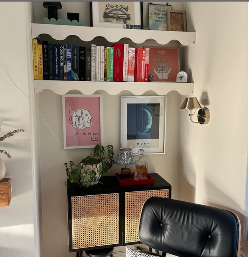 May include: A white shelf with scalloped edges, holding books, artwork, and a small clock. The shelf is mounted on a white wall. Below the shelf is a black cabinet with two doors, each with a woven rattan panel. A black leather chair is in the foreground.