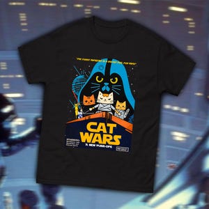 Może przedstawiać: Czarny T-shirt z grafiką kota w hełmie Dartha Vadera trzymającego miecz świetlny. Grafika jest parodią plakatu filmu Star Wars "Przebudzenie Mocy" i zawiera napis "Cat Wars: A New Purr-ope".