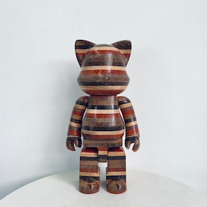 Puede incluir: Una figura de juguete de madera con un patrón a rayas en tonos marrones, rojos y blancos. La figura tiene una cabeza de gato y está de pie sobre una superficie blanca.