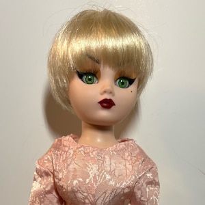 Puede incluir: Una muñeca con cabello rubio y ojos verdes que lleva un vestido rosa con un estampado floral blanco.