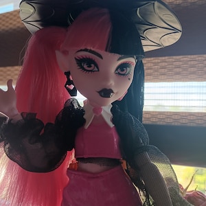 May include: A pink and black doll with a witch hat, wearing a black mesh top with a white collar, a pink crop top, and a pink skirt. The doll has pink and black hair, black eyeliner, and red lipstick.