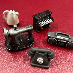 Puede incluir: Un conjunto de artículos domésticos en miniatura, que incluye una máquina de coser negra, una tetera eléctrica blanca, un teléfono de disco negro, una radiocasete negra y un órgano en miniatura negro.