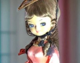 May include: A vintage doll with a pink dress, a brown wig, and a gold-trimmed hat. The doll is wearing a white veil and has blue eyes.