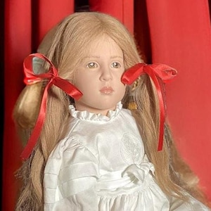 Puede incluir: Una muñeca de porcelana con cabello rubio que lleva un vestido blanco y lazos rojos. La muñeca está sentada en un taburete de madera frente a una cortina roja y azul.