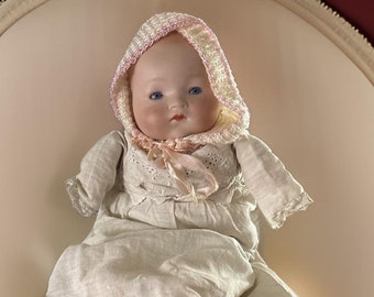 May include: A porcelain baby doll wearing a white dress and a pink and white knitted bonnet. The doll is lying on a white oval-shaped cushion.