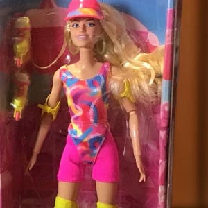 Może przedstawiać: Lalka Barbie w różowo-niebieskim kostiumie kąpielowym, żółtych ochraniaczach na kolana i różowym kapeluszu. Lalka trzyma żółte rolki i jest zapakowana w różowe pudełko z napisem "Barbie The Movie".
