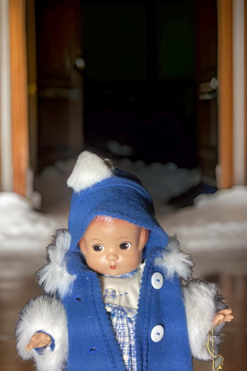 Op de afbeelding: Een porseleinen pop die een blauwe jas draagt met witte bontkraag en een witte hoed. De pop heeft bruine ogen en een geschilderde glimlach.