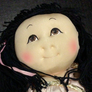 May include: A smiling doll with black hair and pink cheeks. The doll is wearing a pink and gold dress.