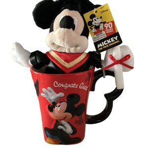 May include: Red ceramic mug featuring a Mickey Mouse design and the text "Congrats Grad". A plush Mickey Mouse wearing a graduation cap and gown is sitting inside the mug. A white plush diploma with a red bow is attached to the mug's handle.