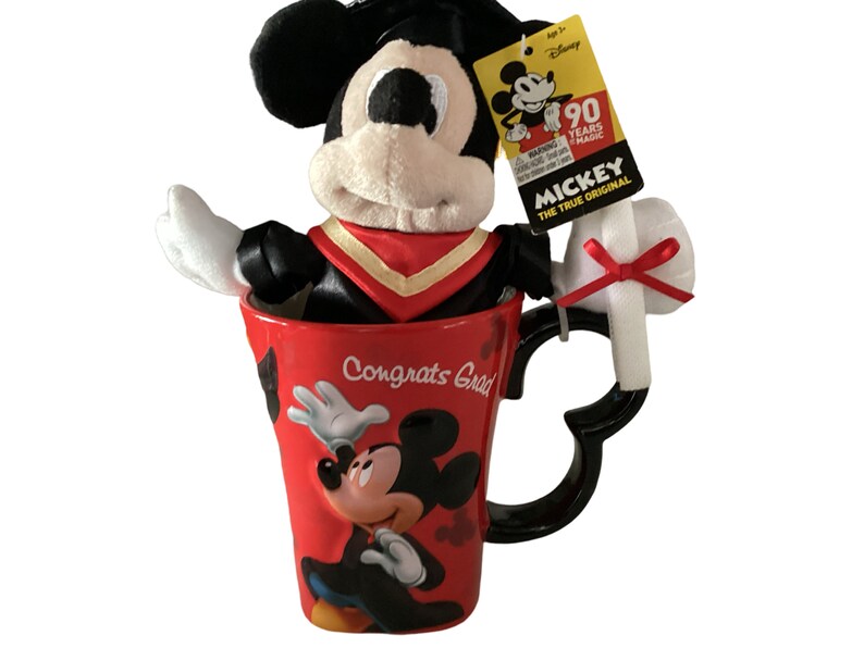 May include: Red ceramic mug featuring a Mickey Mouse design and the text "Congrats Grad". A plush Mickey Mouse wearing a graduation cap and gown is sitting inside the mug. A white plush diploma with a red bow is attached to the mug's handle.