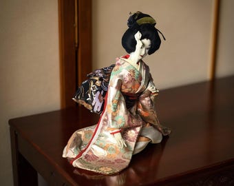 Puede incluir: Muñeca japonesa con kimono tradicional, arrodillada sobre una superficie de madera oscura. El kimono presenta estampados florales en tonos dorados, rosas y verdes. La muñeca tiene cabello negro y una expresión serena. El fondo es de color neutro.
